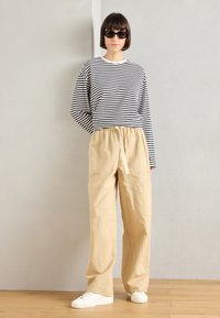 Ριγέ μακρυμάνικη μπλούζα σε μαύρο και άσπρο, συνδυασμένη με ελαφριά μπεζ, φαρδιά παντελόνια με ελαστική μέση, φορεμένα με λευκά αθλητικά παπούτσια.