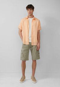 Korte mouwen perzik kleur linnen shirt over een wit T-shirt, gecombineerd met olijfgroene cargoshorts. Bruine sneakers met een gestructureerd oppervlak. Minimalistisch design.