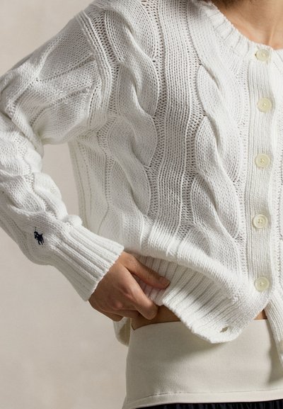 Λευκό πλεκτό ζακέτα με κουμπιά και κεντημένο λογότυπο στην μανσέτα, φορεμένο πάνω από μπεζ ένδυμα, χέρι ακουμπισμένο στη μέση.