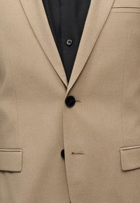 Μπεζ σακάκι με δύο μαύρα κουμπιά και τσέπες με καπάκι, φορεμένο πάνω από ένα μαύρο πουκάμισο με ένα εμφανές κουμπί.