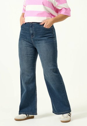 Person trägt weite blaue Jeans, weiße Turnschuhe mit schwarzen Streifen und ein rosa-weiß gestreiftes Hemd, Hände in den Taschen.