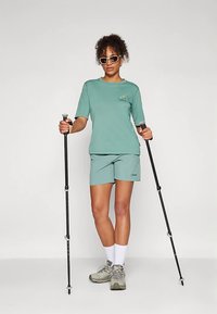 Ανοιχτό πράσινο αθλητικό μπλουζάκι και σορτσάκι από μαλακό ύφασμα. Λευκές κάλτσες, ορειβατικά παπούτσια και ρυθμιζόμενοι μπαστούνια πεζοπορίας ολοκληρώνουν την εμφάνιση.