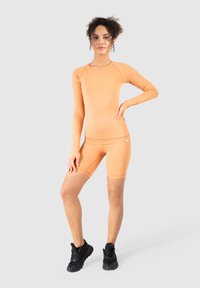 Lyse orange langærmet sports top parret med matchende shorts, der går til midt på låret. Begge dele lavet af strækbart stof; båret med sorte sneakers.