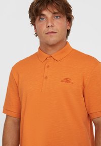 Oranžová polo košeľa vyrobená z textúrovanej látky, s gombíkovou légou a vyšívaným logom tmavšej oranžovej farby na hrudi.