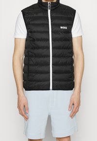 Svart pufferväst med hög krage, framdragkedja och vit logotyp. Bärs över en vit skjorta, ihop med ljusblå shorts.