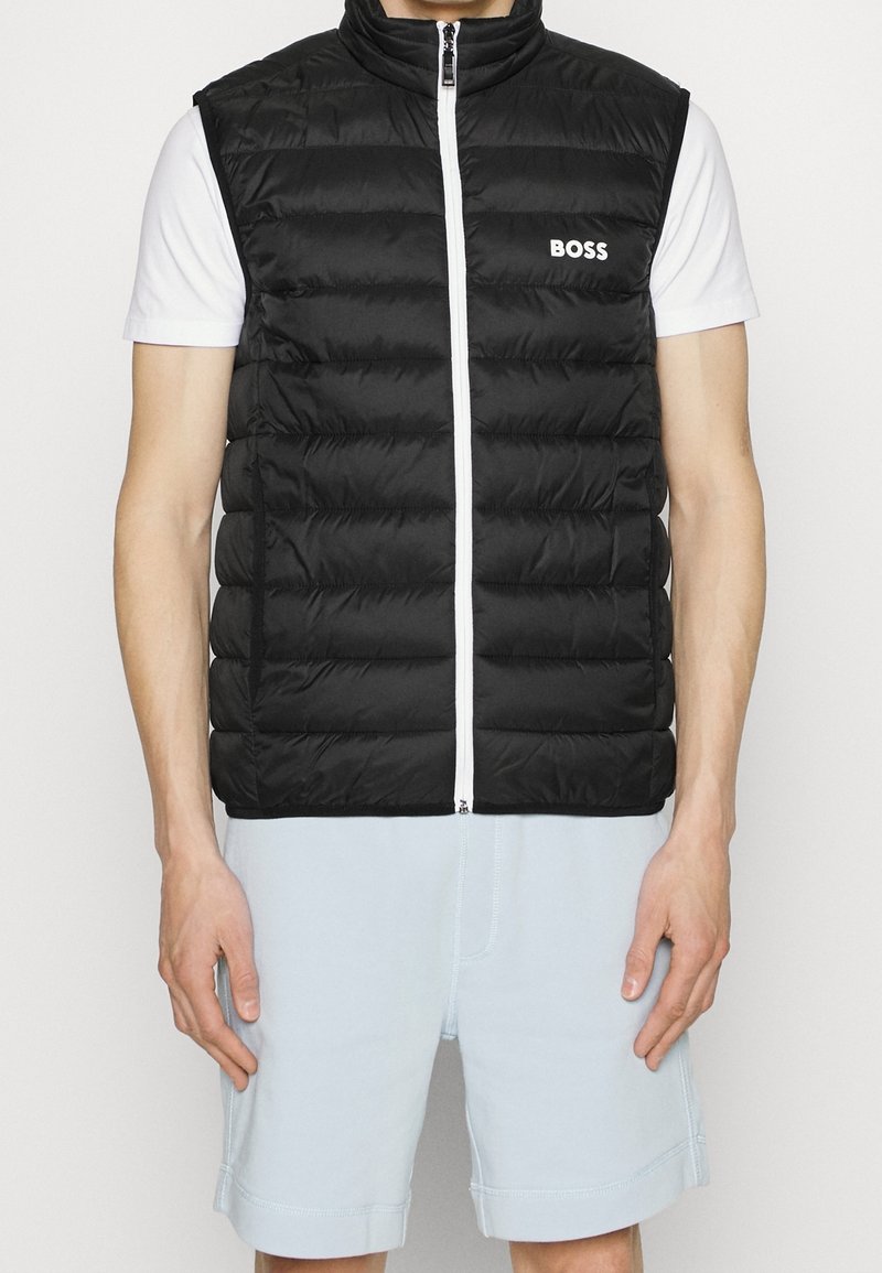 Svart pufferväst med hög krage, framdragkedja och vit logotyp. Bärs över en vit skjorta, ihop med ljusblå shorts.