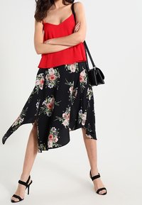 Haut sans manches rouge associé à une jupe asymétrique noire à motifs floraux. Présente des motifs floraux et une texture fluide. Porté avec des talons noirs à lanières.