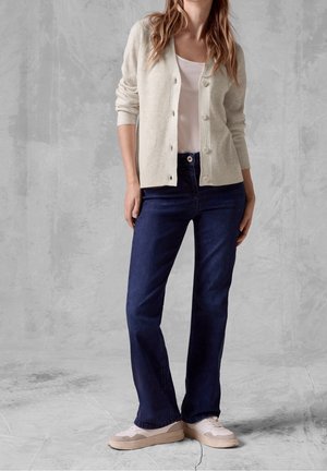 Femme portant un cardigan beige, un haut blanc, un jean bleu foncé et des baskets blanches, se tenant devant un fond texturé gris.