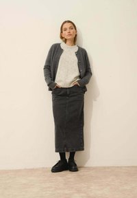 Cardigan gris sobre un suéter jaspeado claro, combinado con una falda maxi de mezclilla oscura y zapatos negros. Diseño simple, detalles mínimos.