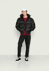 Casaco puffer preto com fecho, gola alta e punhos elásticos, combinado com uma camisa vermelha, calças pretas e ténis pretos com detalhes brancos.
