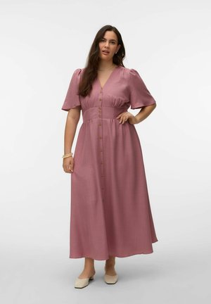 Kvinne med langt mørkt hår som har på seg en knappet lavendel midi-kjole med pufferm og beige sko med lav hæl, stående med en hånd på hoften.