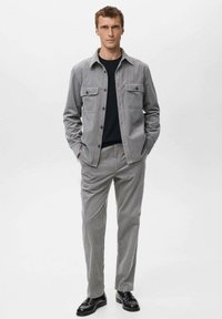 Chaqueta gris de pana con dos bolsillos en el pecho, llevada sobre una camiseta negra de cuello redondo. Pantalones grises a rayas y mocasines negros completan el conjunto.