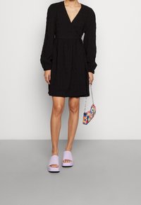 Svart omlotttklänning med långa puffärmar, texturerad tyg och V-ringning. Matchad med lavendelfärgade glidtofflor och en färgglad pärlväska.