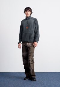 Σκούρο γκρι φλις πουλόβερ με μισό φερμουάρ στον γιακά και μπροστινή τσέπη, συνδυασμένο με καφέ cargo παν trousers που διαθέτουν πολλές πλαϊνές τσέπες και άνετη γραμμή.