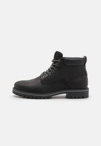 Bota de cuero negro hasta el tobillo con punta redonda, parte frontal con cordones, costura contrastante, tirador y suela de goma resistente con profundos surcos.