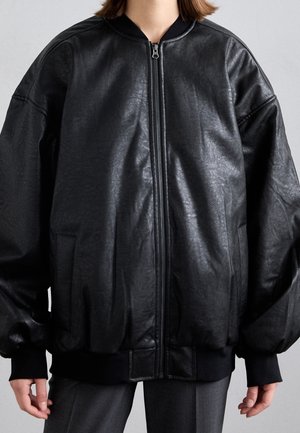 Persoon draagt een oversized zwarte leren jas met ritssluiting, geribbelde boorden en zoom, staand tegen een effen grijze achtergrond.