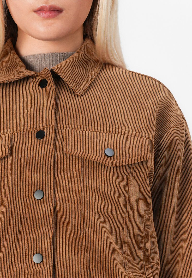 Gros plan sur une personne portant une veste en velours côtelé marron avec des boutons noirs et des poches poitrine sur un col roulé beige à côtes.