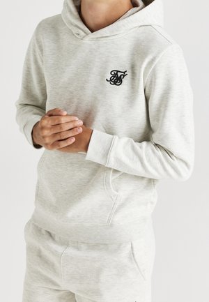 Hombre vestido con sudadera con capucha gris claro y pantalones de chándal a juego, con las manos entrelazadas a la altura de la cintura, logo negro bordado en el pecho, fondo liso.