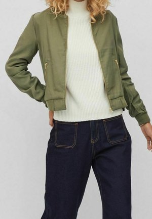 Henkilö, joka pukeutunut oliivinvihreään bomber-takkiin, valkoisen neulotun kauluksellisen villapaidan ja tummansinisten farkkujen ylle, joissa on etutaskut.