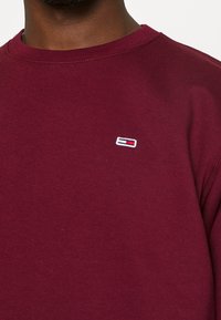 Felpa bordeaux con scollo a costine e logo ricamato in rosso, bianco e blu sul petto. Materiale in cotone, vestibilità casual.