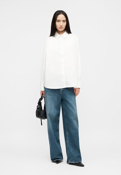 Kvinne med langt mørkt hår iført en løs hvit skjorte, vide blå jeans, svarte spisse sko, holder en liten svart håndveske.