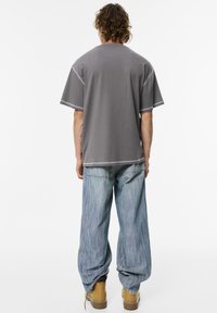 Grå oversized T-shirt med hvid syning, parret med blå brede bukser og gule støvler. Teksturen virker blød og afslappet.