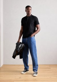 Schwarzes Polohemd mit kurzen Ärmeln, kombiniert mit lockeren blauen Jeans und schwarzen Sneakers. Das Modell hält eine schwarze Lederjacke. Einfaches, lässiges Outfit.