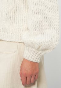 Main de côté portant un pantalon en coton côtelé couleur crème et un pull en laine tricotée épaisse avec une manchette repliée en beige clair.