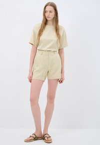 Beige tričkový top s krátkým rukávem zkombinovaný s béžovými šortkami s vysokým pasem a vzorem. Modelka má na sobě hnědé sandály a stojí na bílém pozadí.