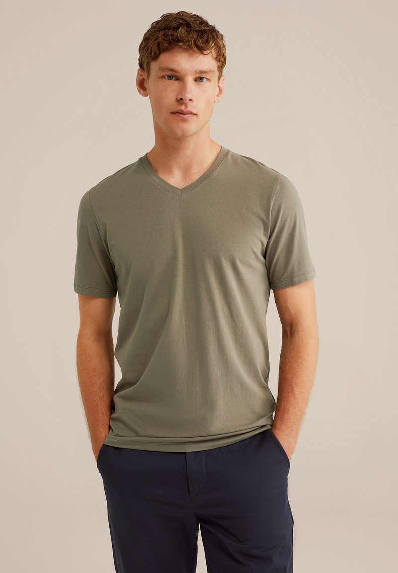 Olajzöld V-nyakú póló, puha anyagból készült, rövid ujjú, egy modellt ábrázol, aki áll, és a kezét a zsebébe dugja.