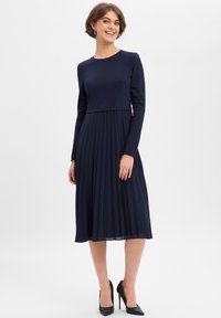 Granatowa sukienka z długim rękawem, dopasowaną górą i plisowaną spódnicą, wykonana z gładkiego materiału. Posiada okrągły dekolt i sięga poniżej kolan.