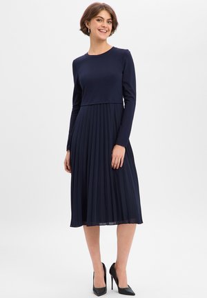 Granatowa sukienka z długim rękawem, dopasowaną górą i plisowaną spódnicą, wykonana z gładkiego materiału. Posiada okrągły dekolt i sięga poniżej kolan.