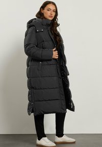 Abrigo acolchado largo negro con capucha, diseño acolchado, cierre frontal con cremallera, bolsillos laterales y forro gris. Combinado con zapatillas blancas.