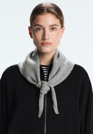 Mlajša ženska s svetlo rjavimi lasmi, spetimi nazaj, nosi črn zgornji del z zadrgo in siv pleten šal, vezan okoli vratu.