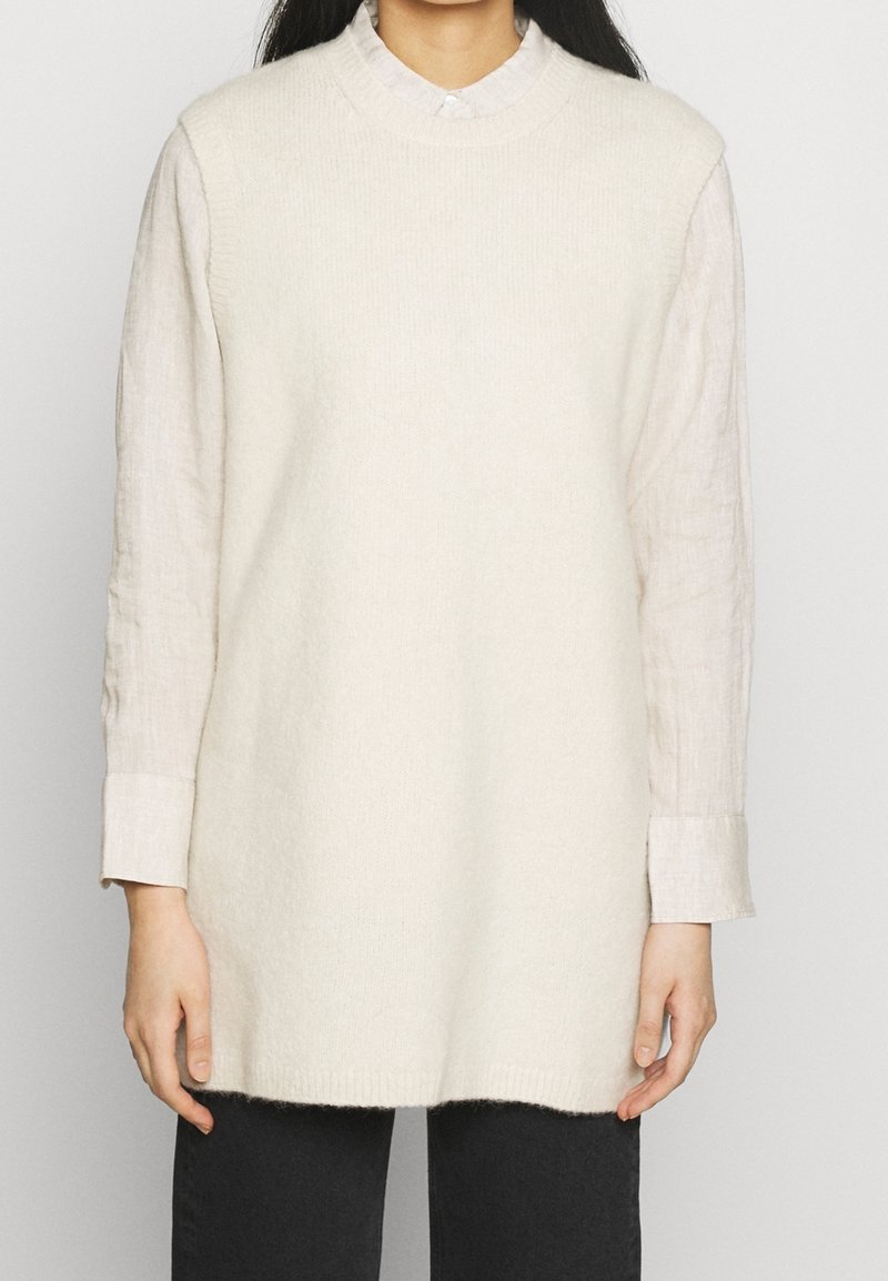 Robe pull en maille crème avec un col haut, superposée sur une chemise boutonnée gris clair, à manches longues et coupe décontractée.