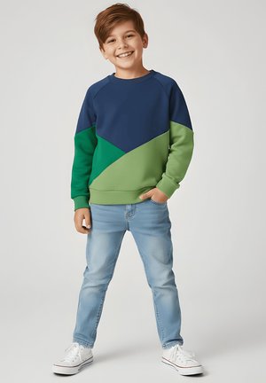 Uśmiechnięty chłopiec stojący z jedną ręką w kieszeni, ubrany w błękitno-zieloną bluzę w kolorowe bloki, jasnoniebieskie dżinsy i białe tenisówki.