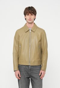 Δερμάτινο μπουφάν ανοιχτό ελιάς με επίπεδη γιακά, κλείσιμο με φερμουάρ και δύο πλαϊνές τσέπες. Ομαλή υφή με εφαρμοστή σχεδίαση, συνδυασμένο με γκρι τζιν.