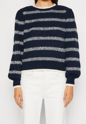 Kobieta ubrana w granatowo-biały sweter w paski z dzianiny z długimi rękawami oraz białe spodnie, stojąca na tle jednolitego białego tła.