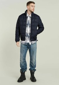 Giacca a vento blu navy con design trapuntato, indossata sopra una camicia a quadrati. Abbinata a jeans sbiaditi e stivali neri con lacci.