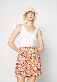Top branco sem mangas combinado com uma saia floral que apresenta flores rosas, laranjas e amarelas sobre um fundo claro. Chapéu de palha bege trançado com detalhes amarelos.