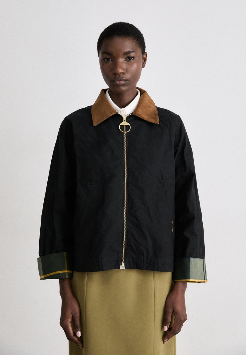Casaco preto com fecho zip, com colarinho de camurça castanho, detalhes em punhos de xadrez verde e um puxador de fecho circular dourado. Apresentado sobre uma roupa bege.