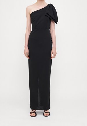 Zwarte lange jurk met één schouder en een grote strikdetail op de schouder, gedragen met zwarte platte sandalen met bandjes, gedragen door een staand model tegen een witte achtergrond.