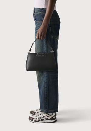 Zwartleren handtas met een korte handgreep, voorzien van een zilveren logo-plaatje. Gecombineerd met losse blauwe denim jeans en bruin-witte sneakers.