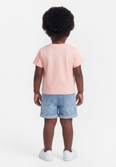 Child with curly hair wearing a light pink t-shirt, rolled-up denim shorts, and white sneakers, standing facing away on a white background.