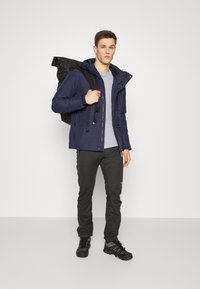 Marineblau isolierte Jacke mit Kapuze, graues T-Shirt, schwarze Hose und robuste schwarze Schuhe. Trägt einen schwarzen Rucksack. Einfaches Design.