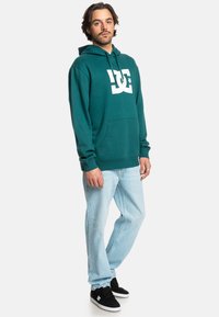 Zelená mikina s bielym logom, v kombinácii so svetlomodrými džínsami a čiernymi topánkami. Mikina má klokanie vrecko a šnúrku na kapucni.