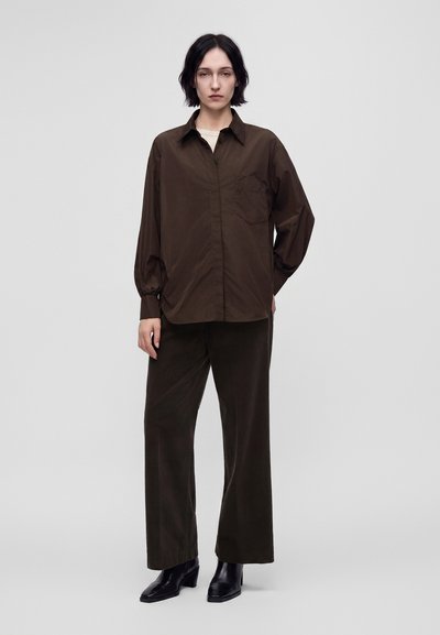 Femme aux cheveux courts et foncés portant une chemise ample marron foncé boutonnée, un pantalon large assorti et des bottines noires sur un fond uni.