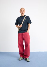 Marineblå bomulls-t-skjorte med grafikk av sky og regndråper, kombinert med løse røde denimbukser. Modell iført grønne sneakers og en beige skulderveske.