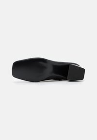 Zapato de tacón negro con un upper suave y una punta ligeramente cuadrada. La suela presenta una textura estriada para ofrecer tracción y estabilidad.