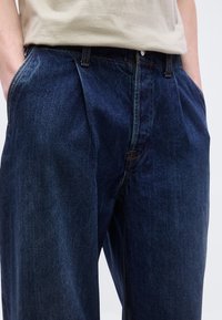 Άτομο φοράει σκουρό μπλε τζιν με πιέτες μπροστά και έχει τα χέρια στην τσέπη, συνδυασμένο με ανοιχτό μπεζ πουκάμισο.